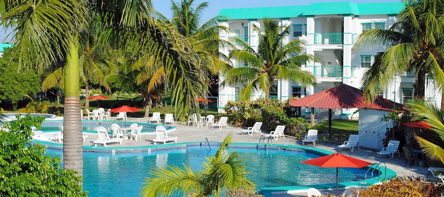 A sunny resort scene with a large turquoise pool, palm trees, patio chairs, umbrellas, and a bright white building with teal accents.