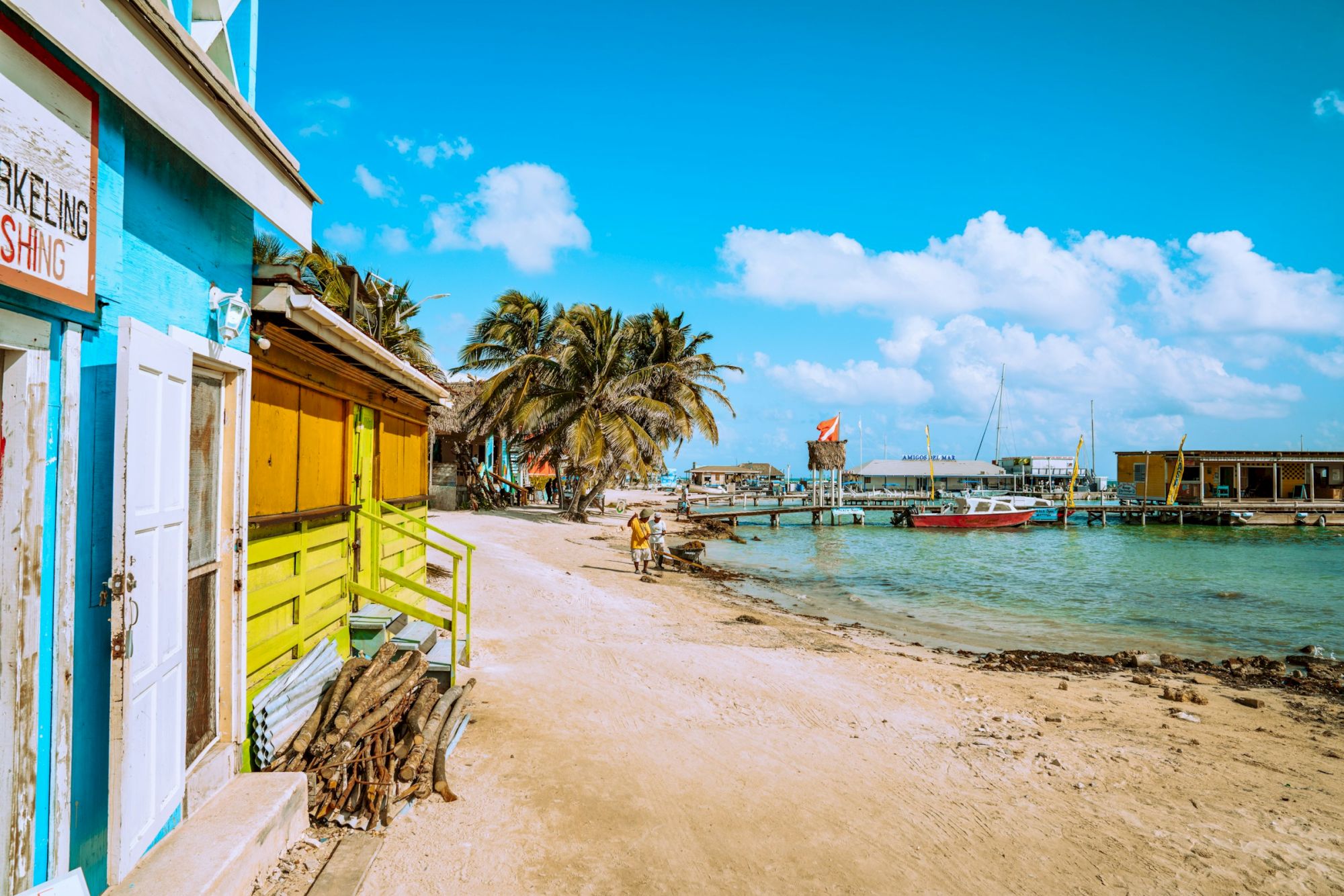 A sunny seaside marina with colorful buildings, palm trees, docked boats, and a calm turquoise shoreline beneath a bright blue sky.