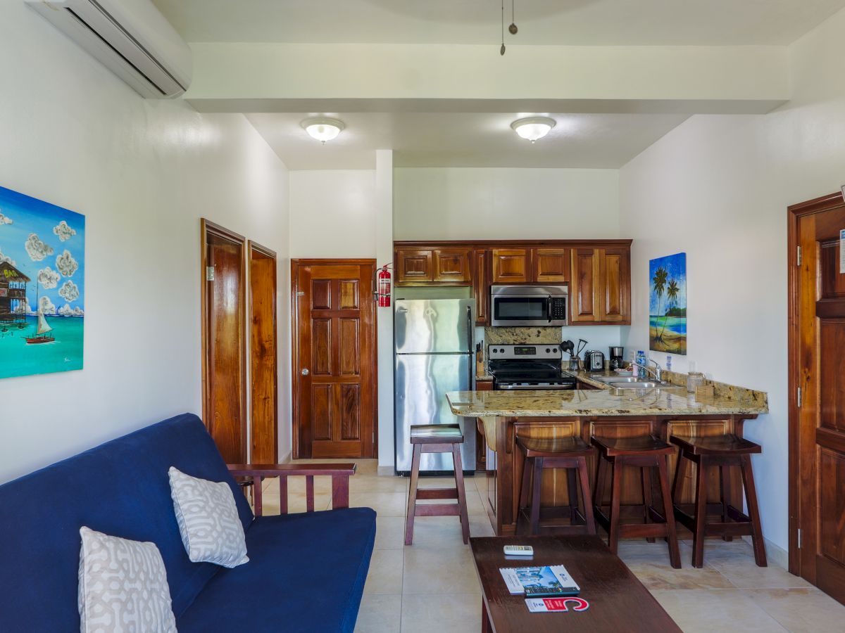 A bright room with a blue couch, kitchen counter, cabinets, fridge, stove, bar stools, and an EXIT door.