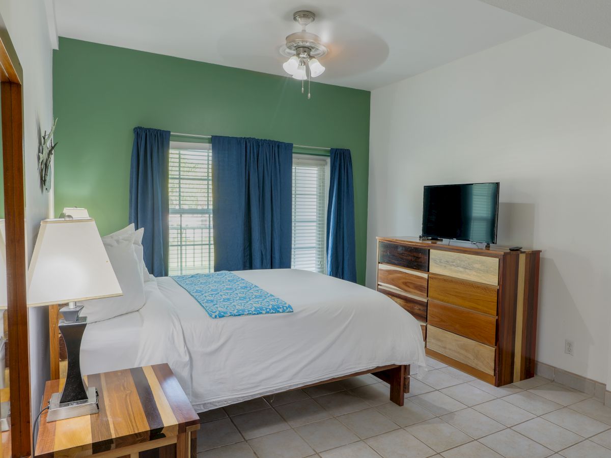 A bright bedroom with a white bed, blue curtains, a ceiling fan, and a TV on a wooden dresser.