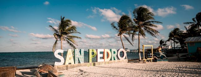 Tropical beach in San Pedro, Belize, with palm trees and a colorful &ldquo;SAN PEDRO BELIZE&rdquo; sign by the ocean.