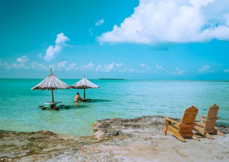 Tropical beach scene: two thatched umbrellas over turquoise water, a person in the sea, and wooden beach chairs on shore.