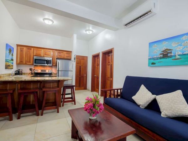 Bright furnished kitchen-living room with wooden cabinets, stove, microwave, blue sofa, and seaside wall art.