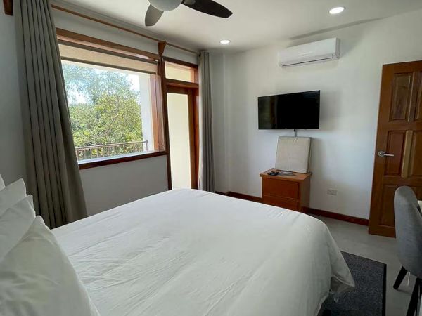 A modern bedroom with a ceiling fan, a neatly made bed, a TV on the wall, and a window with curtains.