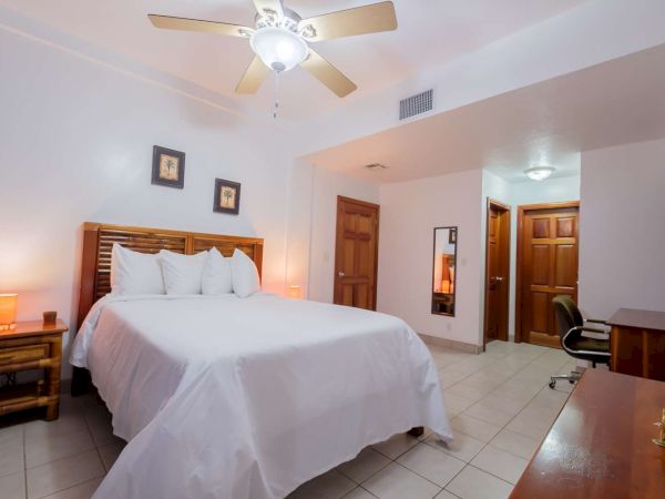 A tidy hotel bedroom with a ceiling fan, neatly made bed, bedside tables, and wood doors along tiled floors.