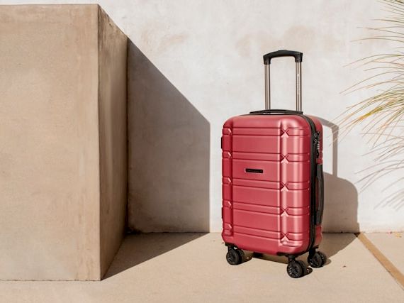 A red hard-shell rolling suitcase stands on wheels in a quiet corner by a wall.