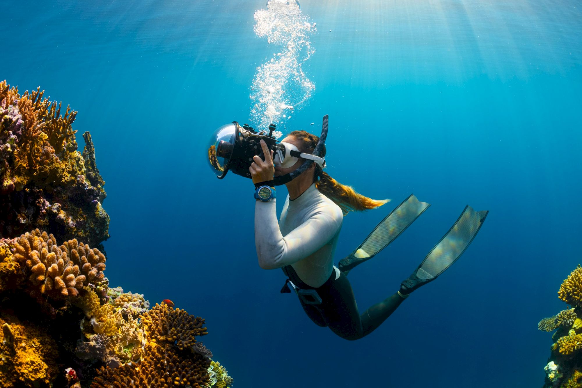 A scuba diver in a mask and fins swims above a coral reef, sending a stream of bubbles into the blue water.