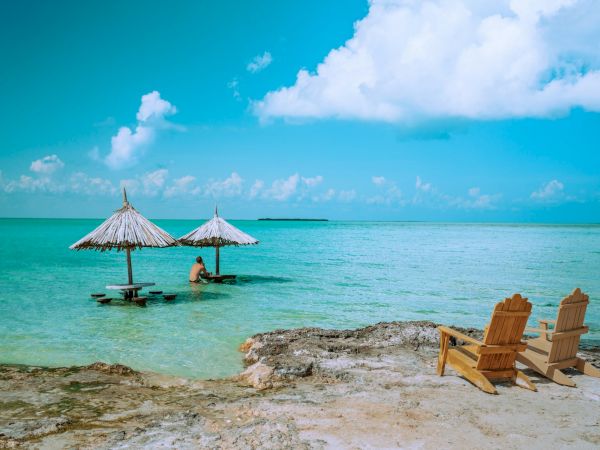 A tropical beach scene with turquoise water, thatched palapa huts and a person relaxing, plus wooden lounge chairs under blue skies.