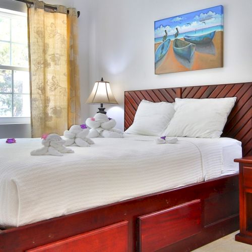 A bright bedroom with a wooden bed, white linens and plush toys, two lamps, seaside wall art, and sheer curtains.