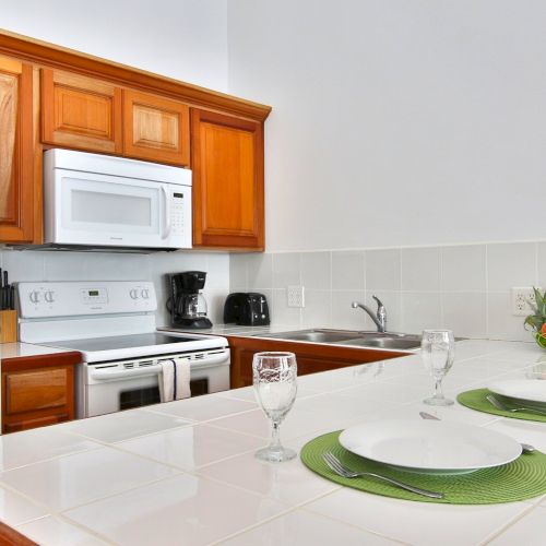 Bright kitchen with wooden cabinets, microwave, stove, sink, and a table set with plates, glasses, and fruit.