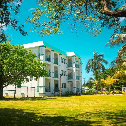 A sunny resort with a white-and-teal multi-story building, surrounded by lush green lawns and palm trees.