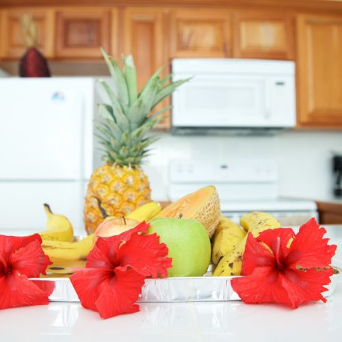 A bright kitchen counter with a fruit bowl&mdash;banana, apple, pineapple&mdash;and red flowers in front.