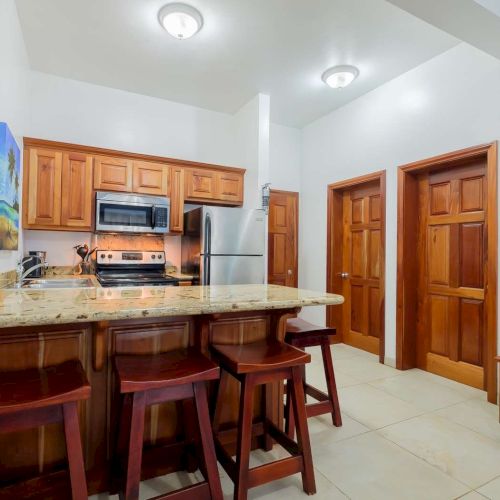 Bright kitchen with wooden cabinets, stove and microwave, a fridge, granite island, bar stools, and two wooden doors.