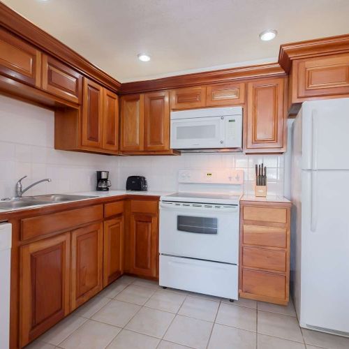 A bright kitchen with wooden cabinets, a white stove and oven, microwave, coffee maker, and a refrigerator under ceiling lights.