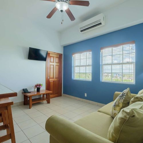 A bright living room with blue accent wall, sofa, lamp, TV, desk, tiled floor, windows with blinds, and a ceiling fan.