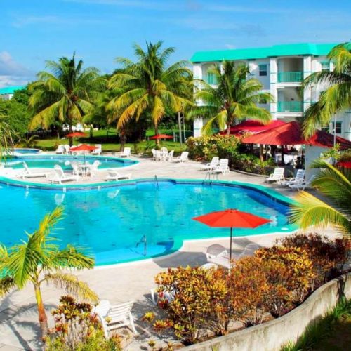 A sunny resort scene with a large turquoise swimming pool, palm trees, red umbrellas, and a white building in the background.