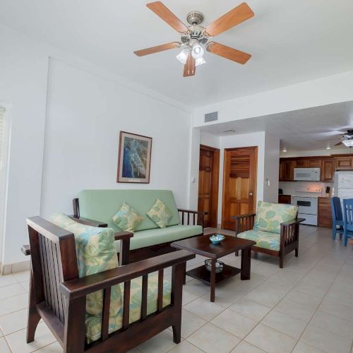 Bright living room with a ceiling fan, tiled floor, and a view into a wooden kitchen with chairs and countertops.