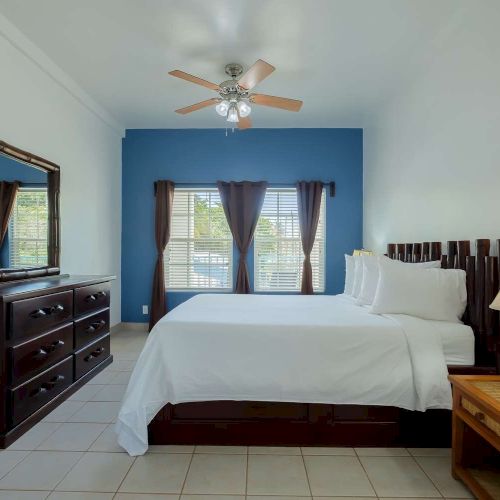 A tidy bedroom with a ceiling fan, double bed, nightstand lamp, chest dresser, and window blinds with dark curtains.