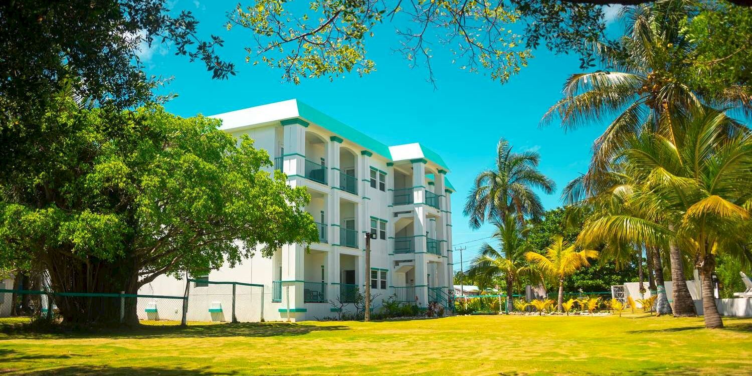 A bright tropical resort with a white-and-teal building, palm trees, and a sunny green lawn under clear blue sky.
