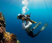 Scuba diver swims above a coral reef, exhaling bubbles in clear blue water.