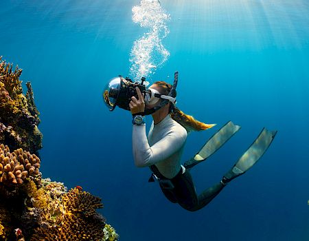Scuba diver swims above a coral reef, exhaling bubbles in clear blue water.