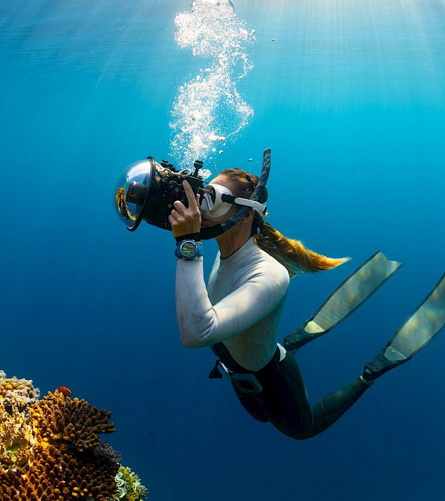 Scuba diver swims above a coral reef, exhaling bubbles in clear blue water.