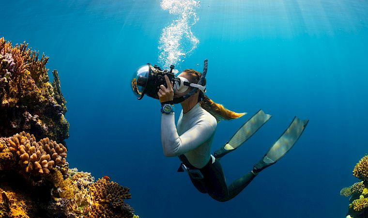 Scuba diver swims above a coral reef, exhaling bubbles in clear blue water.