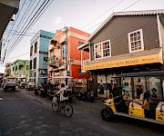 A lively cobblestone street with colorful buildings, parked bikes and carts, and a storefront sign reading “Home of Belize’s Favorite Rums!”.