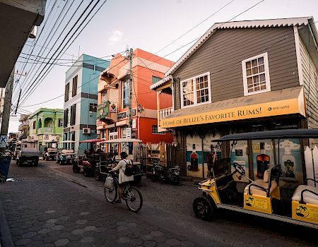 A lively cobblestone street with colorful buildings, parked bikes and carts, and a storefront sign reading “Home of Belize’s Favorite Rums!”.