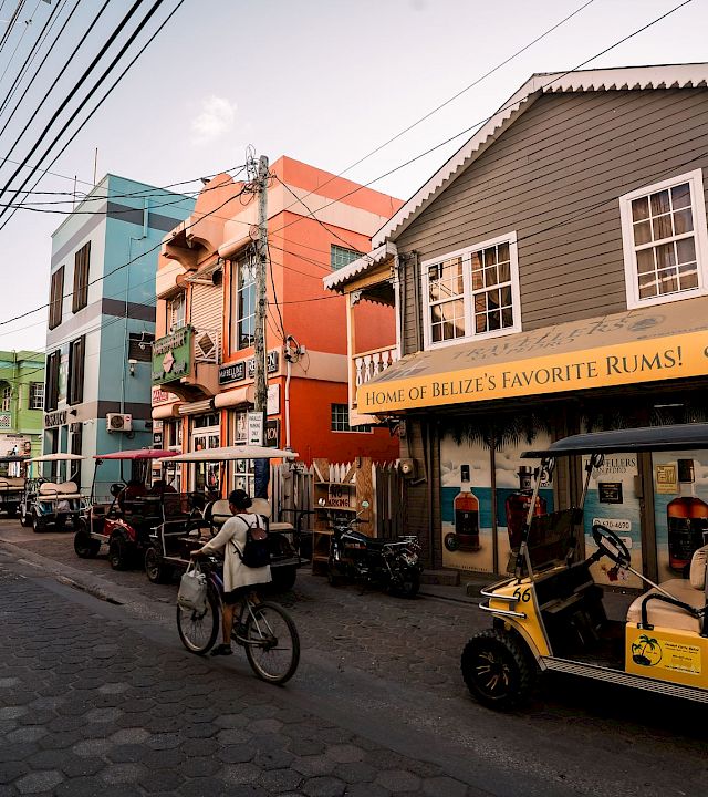 A lively cobblestone street with colorful buildings, parked bikes and carts, and a storefront sign reading “Home of Belize’s Favorite Rums!”.