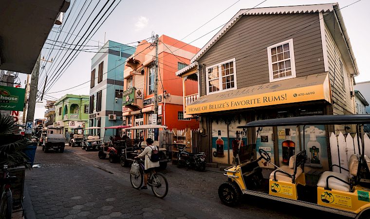 A lively cobblestone street with colorful buildings, parked bikes and carts, and a storefront sign reading “Home of Belize’s Favorite Rums!”.