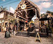 Tropical “PUR” bar with a thatched roof and signs, palm trees, and outdoor seating in sunny weather.