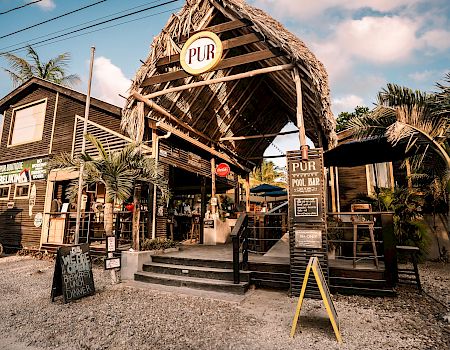 Tropical “PUR” bar with a thatched roof and signs, palm trees, and outdoor seating in sunny weather.