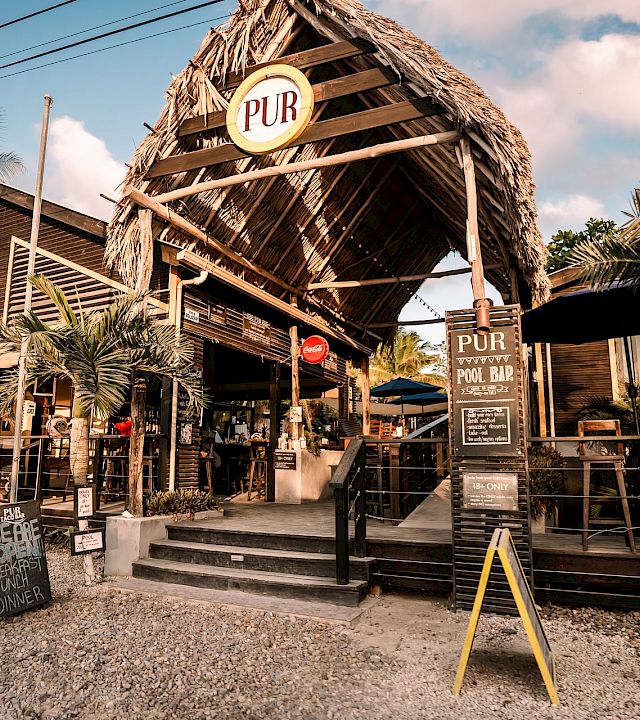 Tropical “PUR” bar with a thatched roof and signs, palm trees, and outdoor seating in sunny weather.