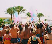 A packed pool party at a sunny beach resort showers the crowd with colorful powder and confetti as everyone cheers.