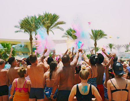 A packed pool party at a sunny beach resort showers the crowd with colorful powder and confetti as everyone cheers.