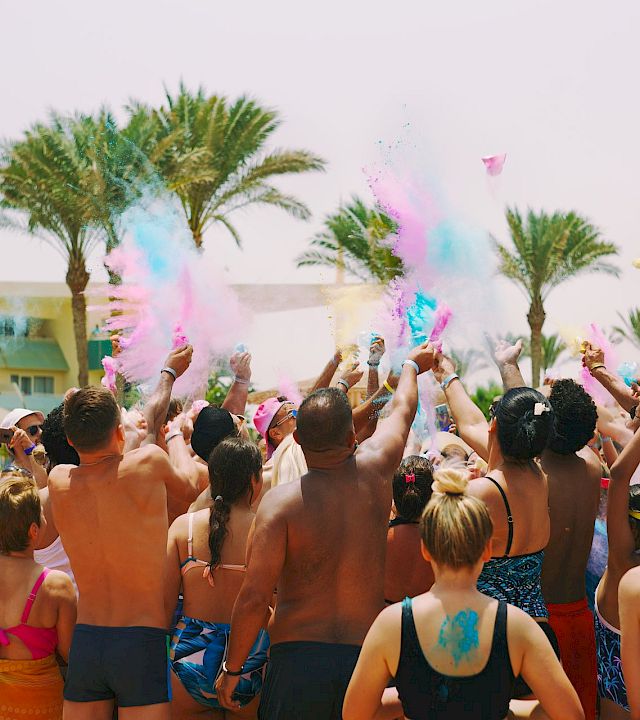 A packed pool party at a sunny beach resort showers the crowd with colorful powder and confetti as everyone cheers.