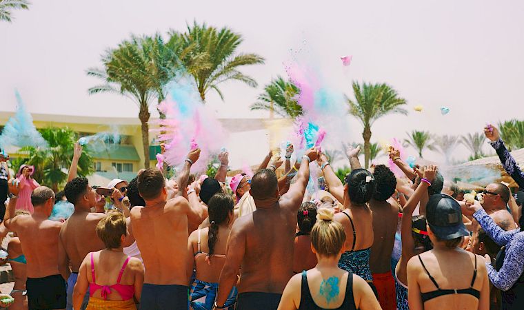 A packed pool party at a sunny beach resort showers the crowd with colorful powder and confetti as everyone cheers.