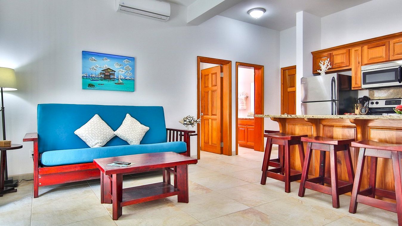 Bright living room with a blue sofa, wooden coffee table, wall art, and an open kitchen with stools, fridge, and microwave.