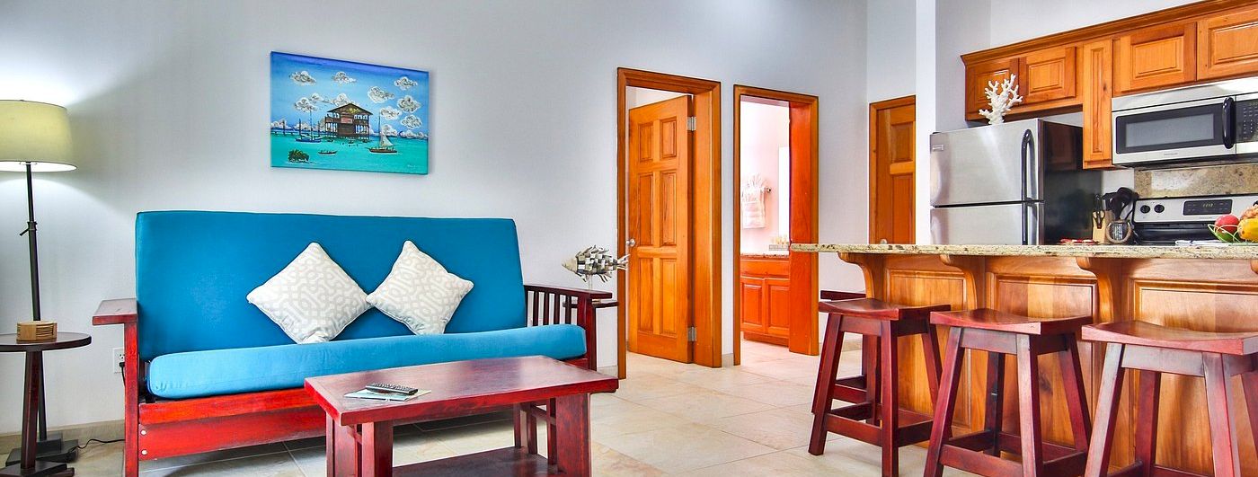 Bright living room with a blue sofa, wooden coffee table, wall art, and an open kitchen with stools, fridge, and microwave.