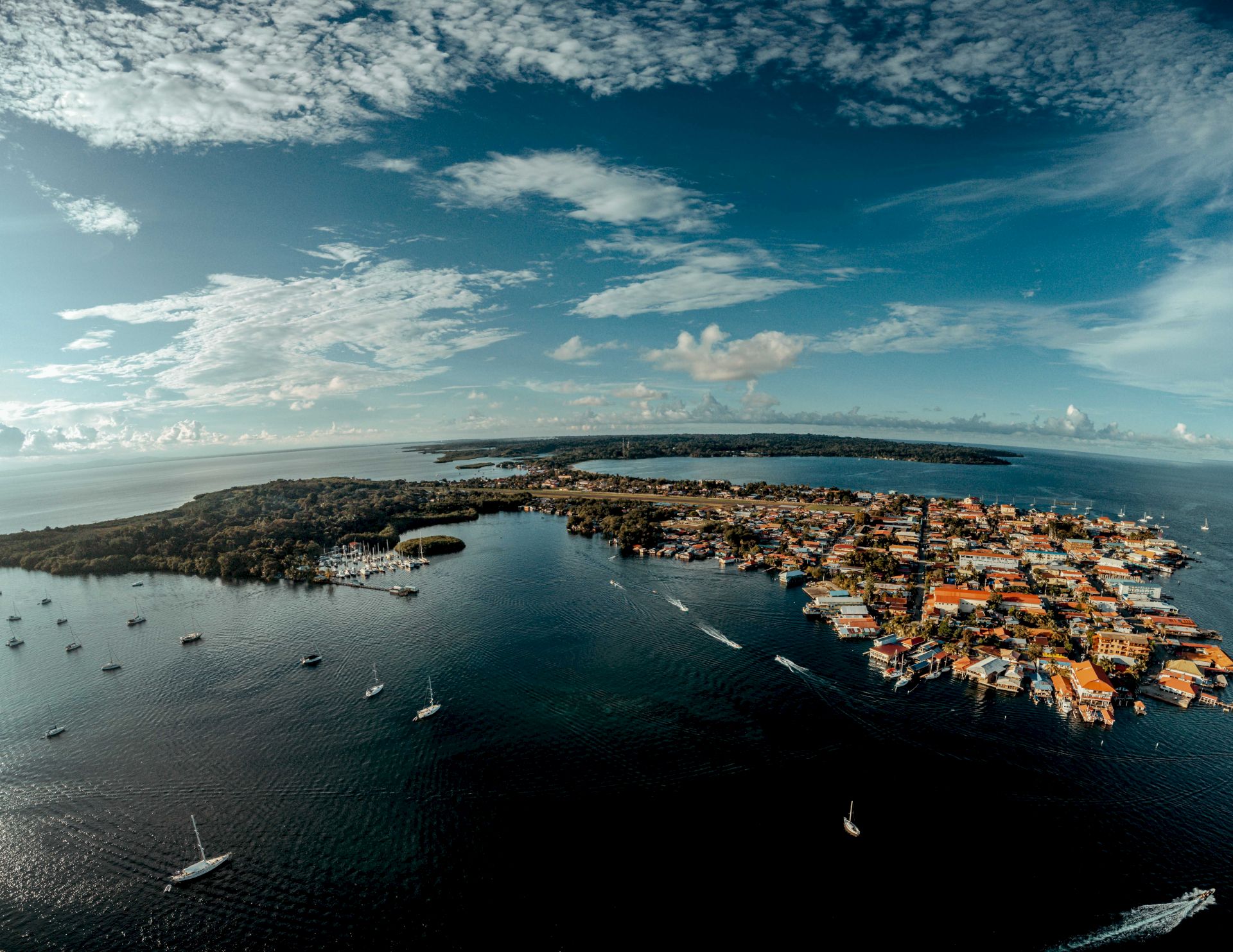 Aerial view of a coastal harbor town with orange-roofed buildings, sailboats and yachts, surrounded by calm blue water and dramatic clouds.