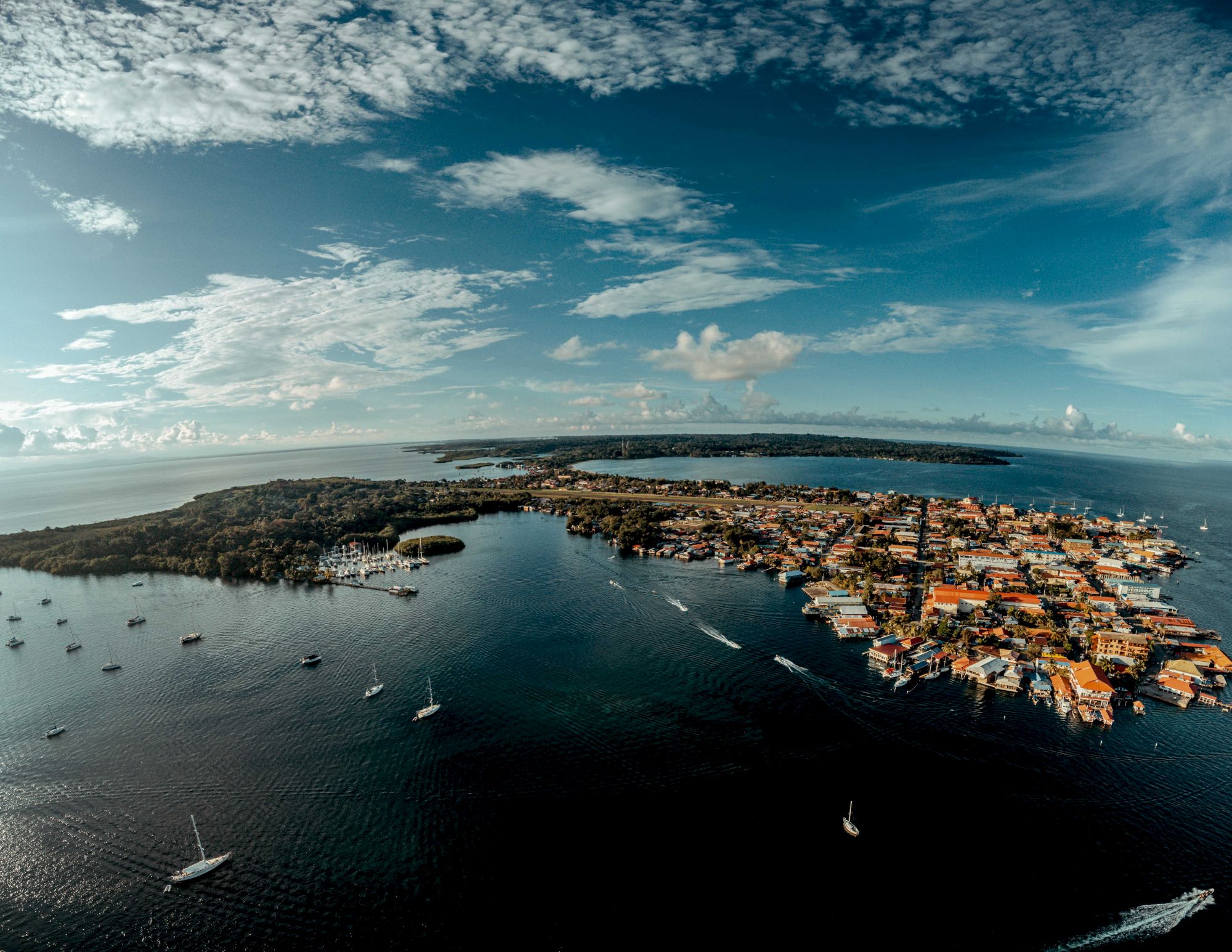 Aerial view of a coastal harbor town with orange-roofed buildings, sailboats and yachts, surrounded by calm blue water and dramatic clouds.