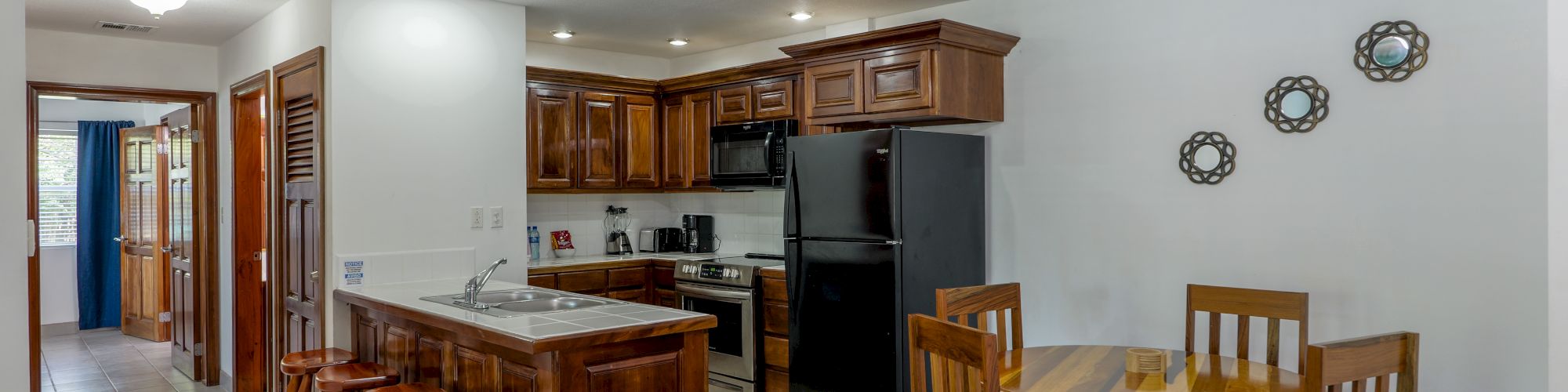 A bright kitchen with wood cabinets, countertop island, black refrigerator and oven, and a dining table under ceiling lights.