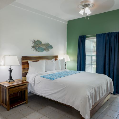 A bright bedroom with a wooden bed, white linens, blue curtains, wall art of fish, and a ceiling fan-light.