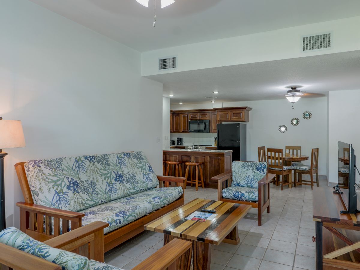 Bright living area with a floral sofa, coffee table, dining set, and a wooden kitchen with appliances and counters.