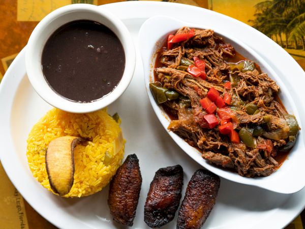A plate of yellow rice with sausage, shredded beef stew with peppers, and a cup of dark soup or sauce.