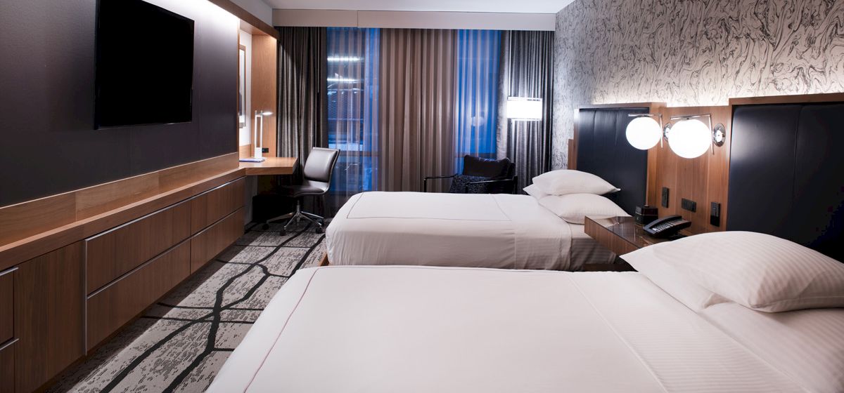 A modern hotel room with two beds, bedside lamps, a TV on the wall, and dark curtains covering large windows.