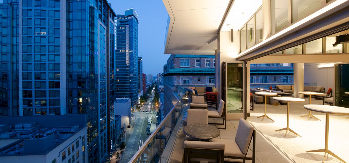 A high-rise balcony with lounge chairs overlooks a downtown street between modern blue office buildings at dusk.